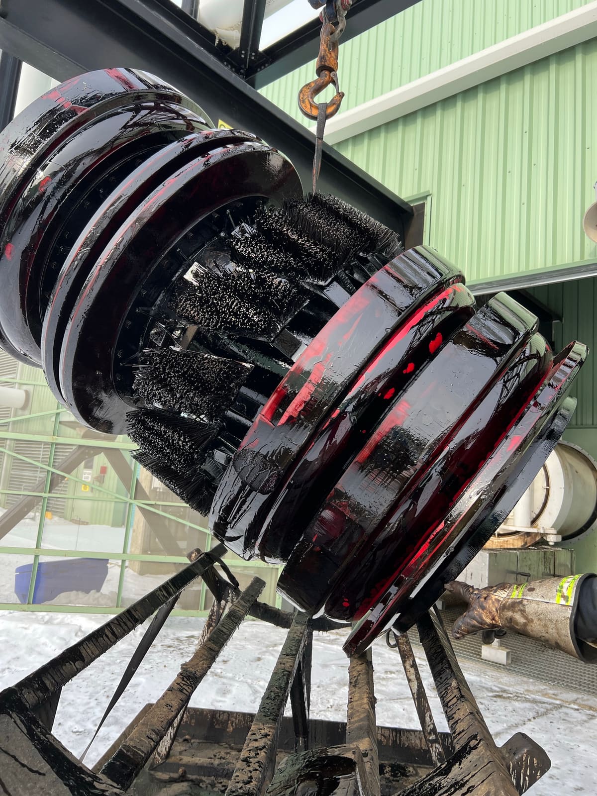 Brush pig coated in heavy bitumen, hanging from a crane hook before cleaning